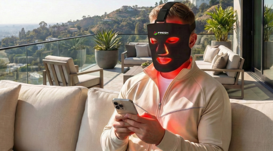 A man using the Fresh Healthcare red light therapy face mask at home to help suport fine lines and blemishes while multitasking on his phone.