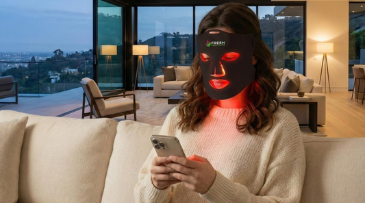 A woman using the Fresh Healthcare red light therapy face mask at home to help support with fine lines and blemishes while multitasking on her phone.